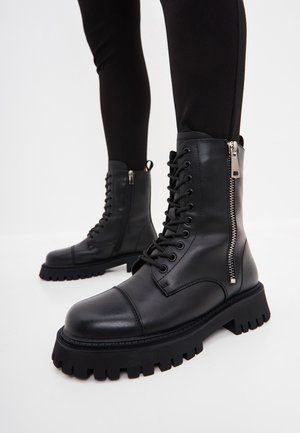 Bottes de combat en cuir noir avec fermetures éclair latérales et semelles épaisses à crampons, portées avec un legging noir sur fond blanc.