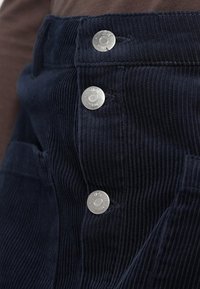 Gros plan d’un pantalon en velours côtelé bleu foncé avec trois boutons argentés visibles étiquetés "JDY DENIM" sur la braguette avant.