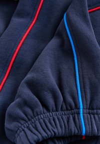 Granatowy sweter z czerwonymi i niebieskimi paskami; cechuje go miękka tekstura i marszczone rękawy, podkreślające sportowe elementy designu.