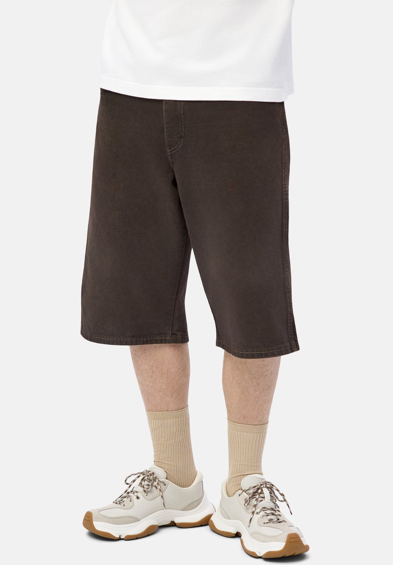 Hombre que lleva pantalones cortos marrón oscuro hasta la rodilla, calcetas beige acanaladas y zapatillas blancas con suelas marrones sobre un fondo claro.