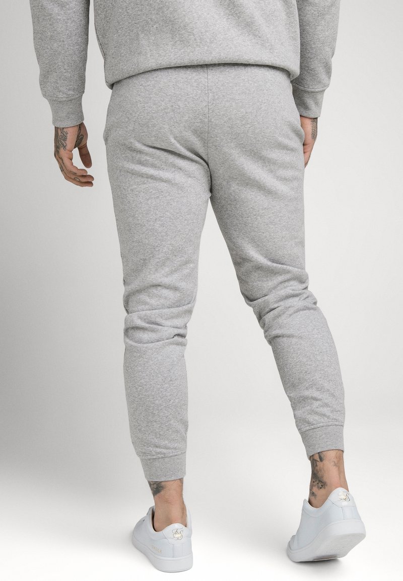 Graue Baumwoll-Jogginghosen mit gerippten Bündchen und elastischem Bund. Getragen mit weißen Sneakers mit goldenen Akzenten. Leichte Textur, körperbetonte Passform.