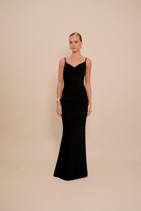 Femeie îmbrăcată într-o rochie de seară fără mâneci, neagră, având cercei aurii și o brățară aurie, stând pe un fundal bej uniform.