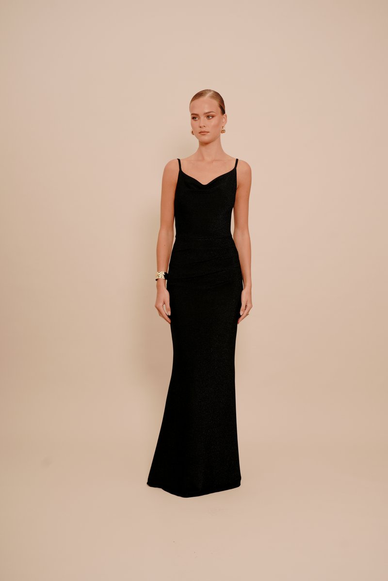 Femeie îmbrăcată într-o rochie de seară fără mâneci, neagră, având cercei aurii și o brățară aurie, stând pe un fundal bej uniform.