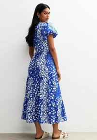 Blaues Blumenkleid mit Puffärmeln, gestuftem Rock und weißen Blumenmotiven. Glattes Material und knöchellanger Schnitt. Getragen mit hellen Sandalen.