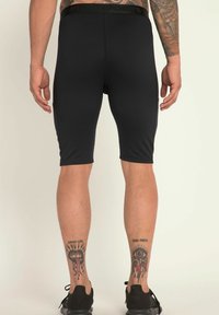 Shorts de sport noirs, coupe ajustée, tissu lisse, longueur jusqu'aux genoux, taille élastique avec logo, portés avec des baskets noires, tatouages visibles sur les mollets.