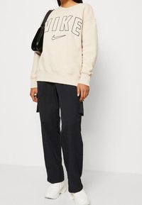 Sudadera beige de Nike con un logo negro audaz, combinada con pantalones cargo azul marino y zapatillas blancas de suela gruesa. Diseño sencillo, corte casual.