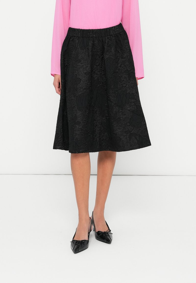Svart midi kjol med texturerat blommönster, elastisk midja och en flödande silhuett. Matchad med spetsiga svarta skor och en rosa topp.