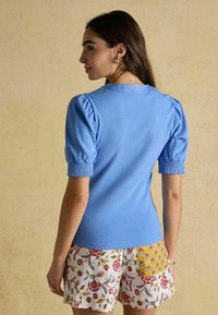 Sieviete ar īsām piedurknēm, zilā ribota augšpusē ar puffed pleciem, un ziedotu šortu ar dzeltenu ziedu kabatu, stāv sānis pret gaiši brūnu sienu.