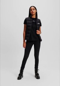 Tânără îmbrăcată cu o vestă pufină neagră, un tricou negru cu logo-ul "Karl", pantaloni skinny negri și cizme negre cu șiret stând în fața unui fundal simplu.