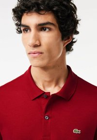 Rød polo-skjorte lavet af tekstureret bomuld, med en klassisk krave, to knapper og et grønt broderet krokodillelogo.