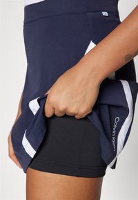 Calvin Klein atlétikai szoknya sötétkék színben, fehér kiegészítőkkel, belső fekete sorttal és sima, rugalmas anyagú textúrával.