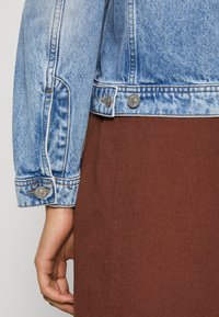 Veste en denim bleu clair avec une texture usée, dotée de poignets boutonnés et de coutures orange, assortie à une jupe marron.