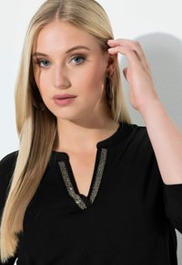 Black top with a V-neck and metallic-accented stitching. Smooth fabric, long sleeves, and lightweight design. Model's hand touches hair.