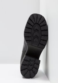 Semelle de botte noire avec un motif de crampons profonds appuyée contre une surface d'angle blanche, affichant la marque "Vagabond" et la taille 37.