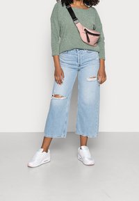 Grüner strukturierter Pullover, hellblaue weit geschnittene Jeans mit ausgefransten Details, weiße Sneakers mit rosa Akzenten und eine rosa Umhängetasche.