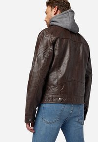 Braune Lederjacke mit grauem Kapuzen, versehen mit einer strukturierten Oberfläche, Druckknopf-Details und einem mittellangen Design, kombiniert mit blauen Jeans.
