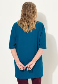 Türkisenes Oversized-T-Shirt mit Ellbogenlängenärmeln, rundem Ausschnitt und glatter Textur. Das Model hat lange, wellige Haare und hält die Hände hinter dem Rücken.