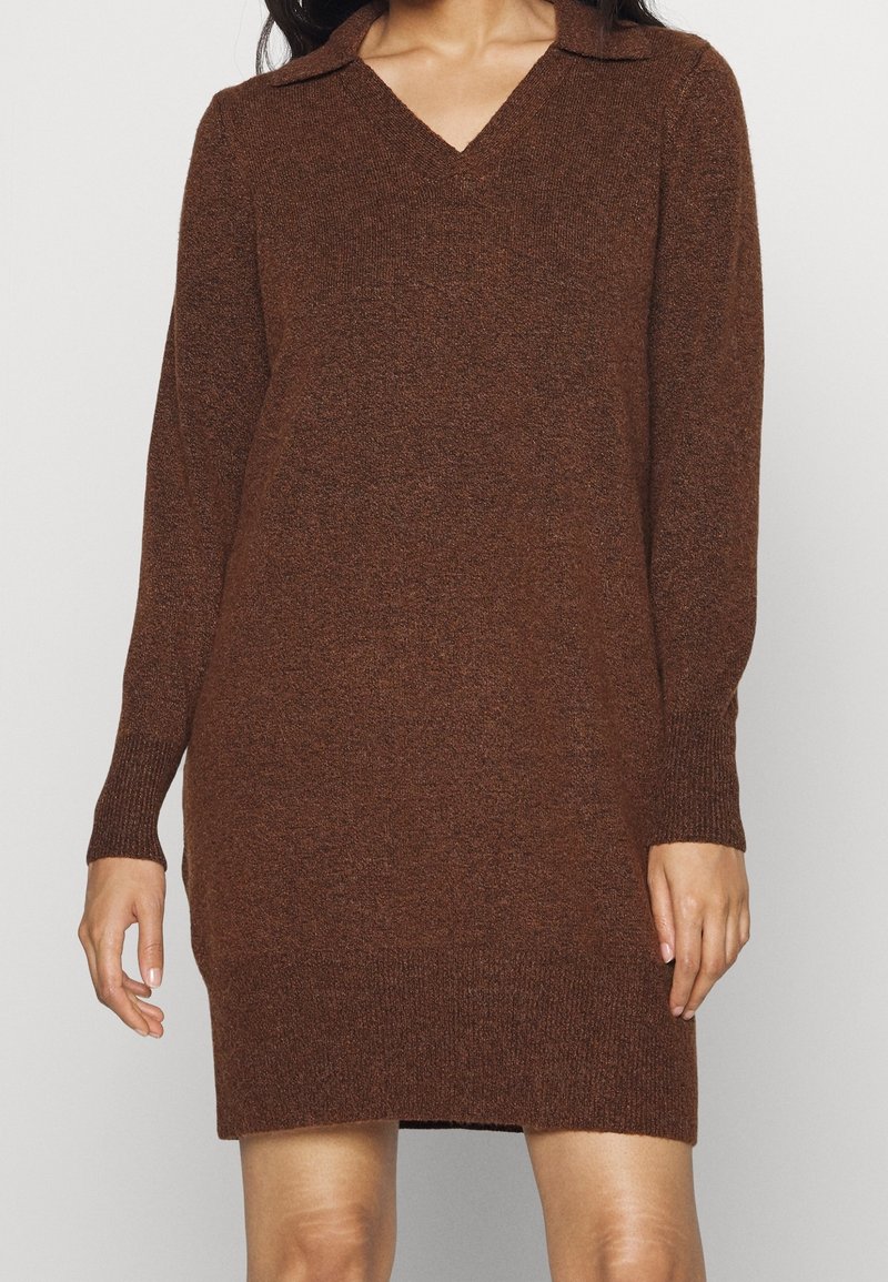 Femme portant une robe pull en maille brune texturée à manches longues avec un col en V, debout devant un fond uni.