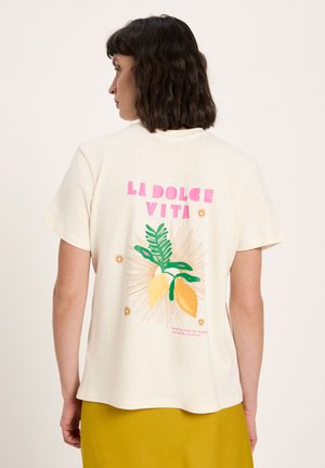 Woman with dark hair wearing cream t-shirt with pink "LA DOLCE VITA" text and lemon and leaves graphic on back, paired with mustard skirt.
