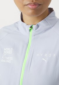 Lys grå zip-up jakke med høj krave. Har en neon grøn lynlås og hvid tekst. Materialet virker glat med et geometrisk mønster.