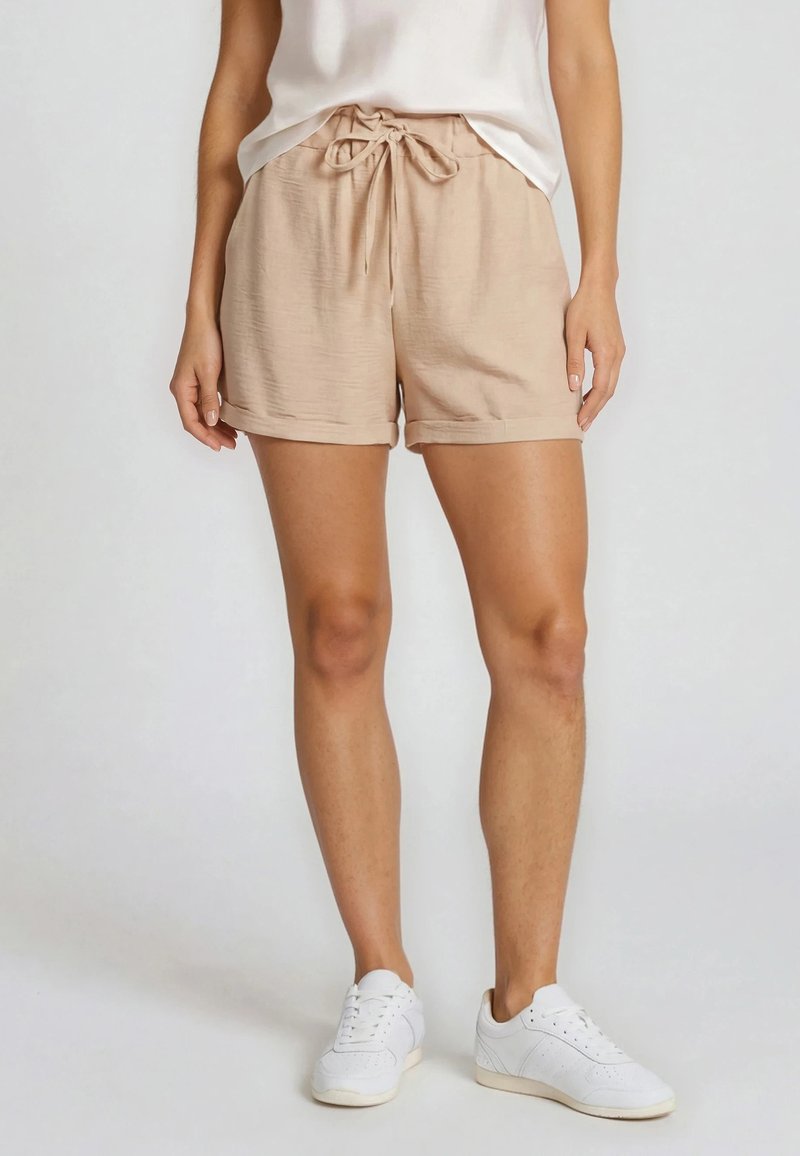 Femme portant un short beige à cordon, un haut sans manches blanc et des baskets blanches sur un fond clair uni.