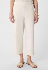 Cremefarbene weite Hose aus glattem Stoff mit hoher Taille. Kombiniert mit beigen, gewebten Slippers.