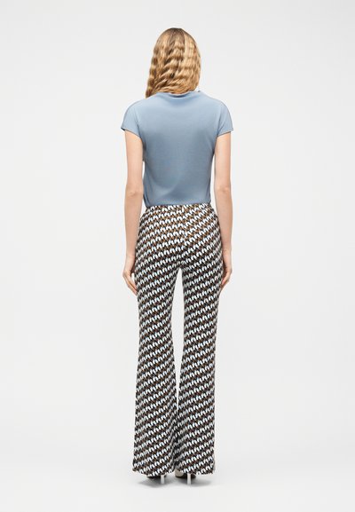 Γυναίκα με μακριά κυματιστά ξανθά μαλλιά στέκεται γυρισμένη προς τα πίσω, φορώντας μπλε πουκάμισο με κοντά μανίκια και φαρδιά παντελόνια με σχέδια, συνδυασμένα με ψηλοτάκουνα.