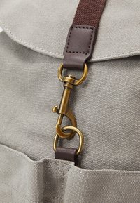Mochila de lona gris con una correa de cuero marrón y herrajes de latón, que presenta una superficie texturizada y un bolsillo frontal.