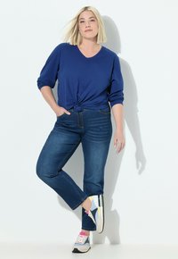 Blaues Langarmoberteil mit geknoteter Front, kombiniert mit dunkelblauen Jeans und mehrfarbigen Sneakers mit strukturiertem Sohlen und kontrastierenden Akzenten.