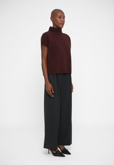 Mujer con un suéter sin mangas color burdeos, pantalones de pinza negros de pierna ancha y tacones negros de punta, de pie de lado.