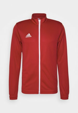 Chaqueta deportiva roja con mangas largas, cuello alto, cremallera blanca de longitud completa y logo blanco de Adidas en el lado izquierdo del pecho. Textura de tela suave.
