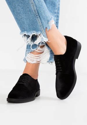 Des chaussures en velours noir à lacets portées avec un jean en denim bleu clair déchiré sur un fond blanc.