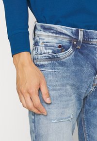 Světlé modré džínové kalhoty s vybledlým vzhledem, opotřebovanými detaily a značkovou nášivkou na zadním kapse. Nošeno s modrým dlouhým rukávem.