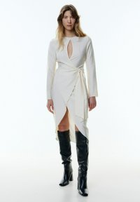 Helle beige Wickelkleid mit langen Ärmeln, einem Schlüsselloch-Ausschnitt und Schlitzsaum, kombiniert mit knielangen schwarzen Lederstiefeln. Glatte Textur.
