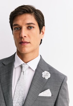 Junger Mann mit grünen Augen, der einen grauen Anzug, eine weiße Krawatte mit floralem Muster, ein weißes Hemd und eine weiße Blütenanstecknadel vor einem weißen Hintergrund trägt.