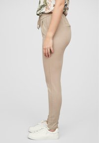 Beige joggebukser med snøring i midjen og sidelommer, laget av myk stoff, vist fra siden med hvite sneakers.