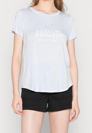 Lyseblå kortærmet T-shirt med teksten "Hollister California" og fuglelogo, parret med sorte shorts på en stående person.