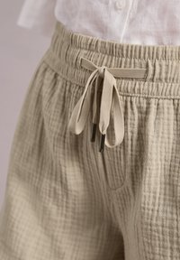 Pantalon beige texturé avec taille élastique et cordon noué, porté avec une chemise blanche boutonnée rentrée dans le pantalon.
