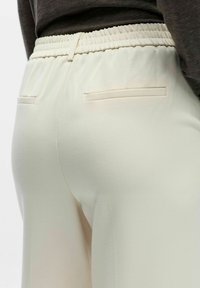 Lyse beige elastiske bukser med glatt tekstur, to baklommer og en avslappet passform.