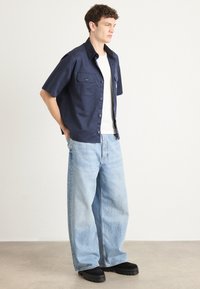 Marinblå kortermet skjorte med to frontlommer, sammen med baggy lyseblå denimjeans og svarte sko med tykk såle.