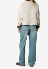 Person iført en beige jakke med brune manchetter, løstsiddende blå jeans og mørke sko, stående med ryggen til betrageren.