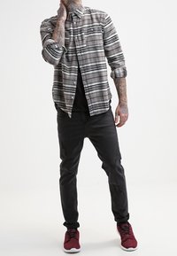 Camicia di flanella a scacchi grigia e nera, colletto con bottoni, maniche lunghe, abbinata a jeans neri strappati e scarpe da ginnastica rosse.