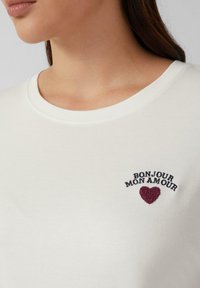 Weiße kurzärmlige Bluse mit Rundhalsausschnitt, verziert mit burgunderrotem gesticktem Text "BONJOUR MON AMOUR" und einem kleinen Herzdetail. Glatte Textur.
