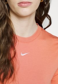 Žena oblečená v oranžovém tričku Nike s bílým logem swoosh u límce a zlatými kruhovými náušnicemi, s volnými hnědými vlasy.