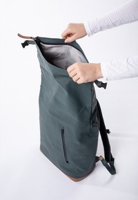 Grüner wasserdichter Rucksack mit glatter Oberfläche, Reißverschlussverschluss und braunen Lederakzenten. Das Innere weist ein gittermusteres Futter auf.