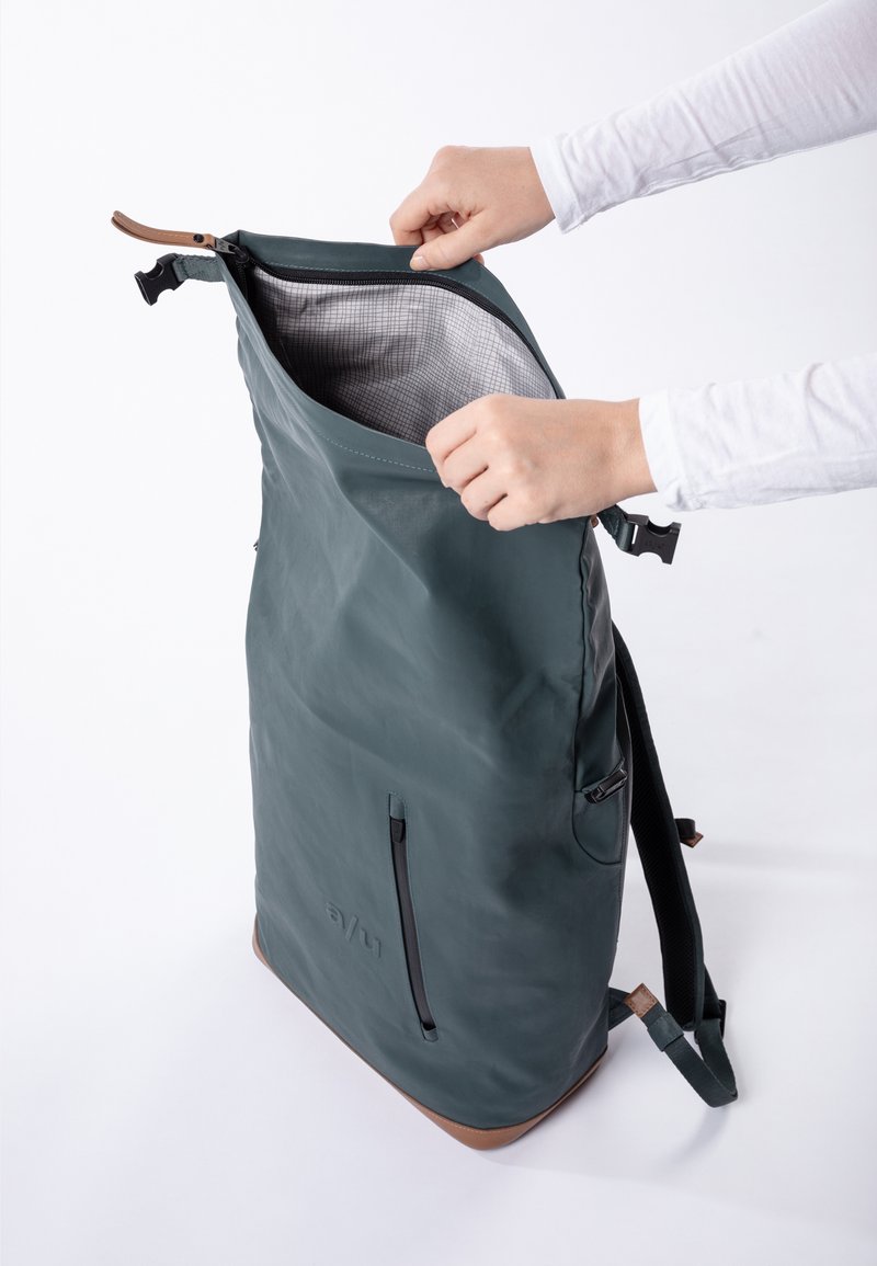 Grüner wasserdichter Rucksack mit Rollverschluss, innen grau kariert und braunen Lederakzenten. Ausgestattet mit verstellbaren Trägern und Seitentasche.