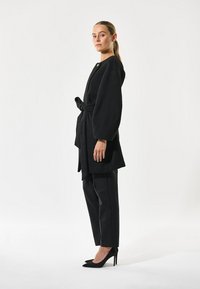 Schwarzer, weicher Stoffmantel mit einem Bindegürtel, langen Puffärmeln und einem einzelnen goldfarbenen Knopfakzent, kombiniert mit schwarzen maßgeschneiderten Hosen und High Heels.