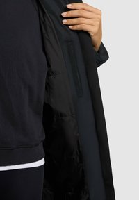 Zwarte jas van glad, gewatteerd doek; heeft een zichtbare voering, zijzak en is iets open, waardoor een donker topje eronder zichtbaar is.