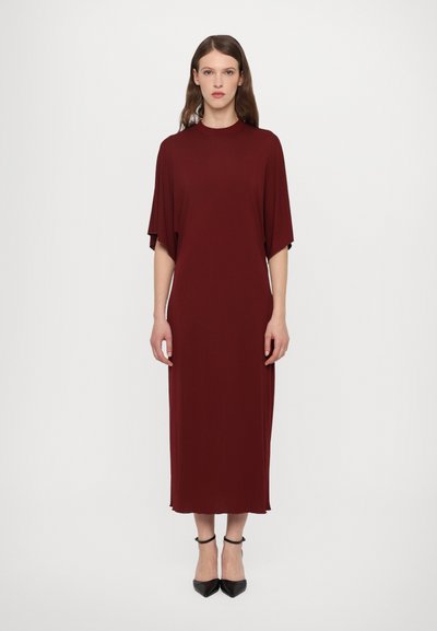 Robe maxi bordeaux avec un col rond, des manches courtes, une texture douce et une silhouette droite, assortie à des escarpins noirs à bout pointu.
