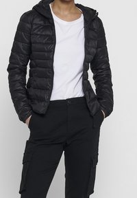 Black, puffy jacket with a zippered front and hood, featuring a quilted design and elastic cuffs. Worn over a white shirt and black pants.